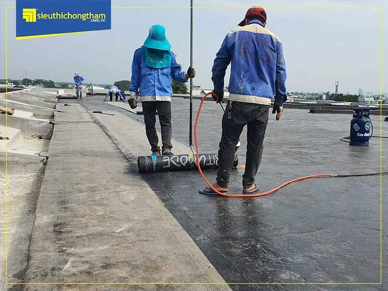 Người thợ đang thi công chống thấm nhà vệ sinh bằng màng khò nóng, sử dụng đèn khò để làm nóng chảy và dán màng bitum chắc chắn lên bề mặt sàn.